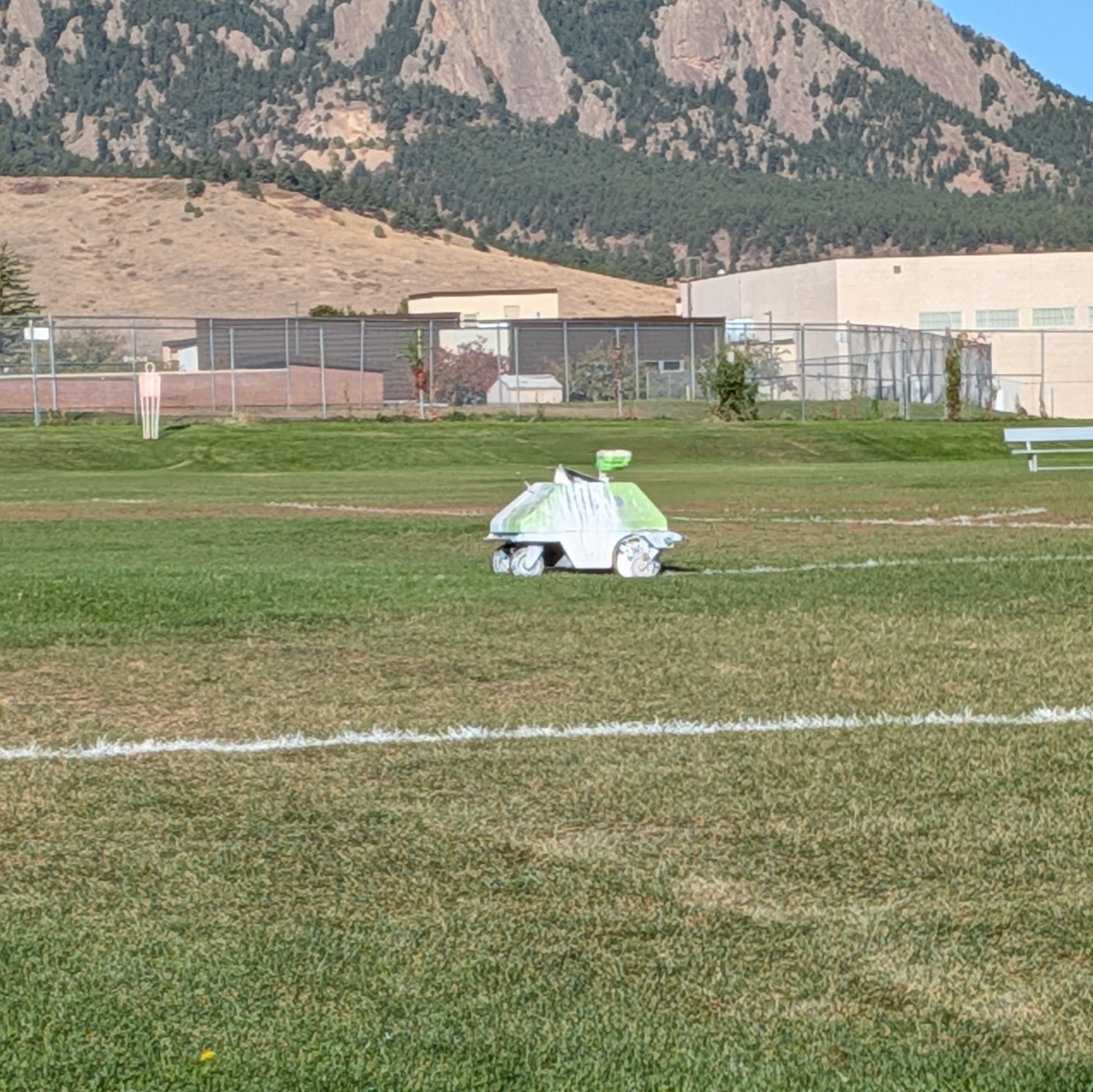A small white and green robot painting white boundary lines on a grass field at Fairview High School, with the Flatirons behind